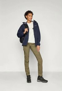 Sieviete stāv ar tumši zilā žaketē, balto kreklu, olīvzaļām kravas biksēm, melnām pārgājienu zābakiem un nēsā melnu mugursomu uz viena pleca.