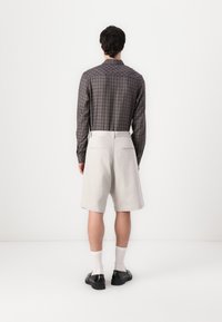 Rutet langærmet skjorte i mørkegrå og brun, parret med lysebeige shorts, hvide sokker og sorte sko. Minimalistisk design.