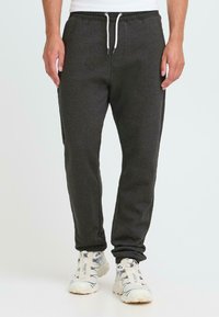 Mann iført mørkegrå joggebukser med hvit snor og hvite joggesko, som står mot en lys bakgrunn.