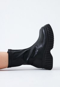 Botas negras hasta la rodilla hechas de un material brillante y suave, con una suela de plataforma gruesa y estriada. Diseño elegante con una forma ajustada.