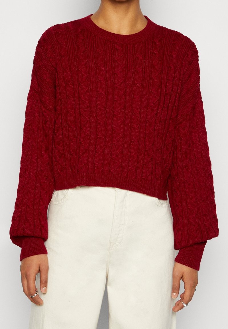 Personne portant un pull à torsades rouge foncé à manches longues et un pantalon taille haute couleur crème, debout avec les mains relâchées le long du corps.