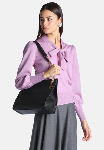 Femme portant un pull rose avec un nœud et une jupe grise tenant un grand sac à main noir avec une bandoulière chaîne dorée sur son épaule.