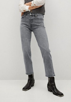 Osoba nosząca wyblakłe szare dżinsy z wysokim stanem i czarne botki za kostkę na suwak, stojąca ze skrzyżowanymi ramionami na jednolitym jasnym tle.