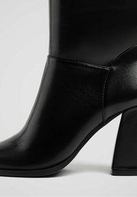 Bottine en cuir noir avec un talon épais, texture lisse et un design courbé au niveau de la cheville. Détails de couture sur le côté visibles.