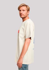 Cream-colored oversized cotton t-shirt with short sleeves, featuring red and blue text and graphic accents on the front and back.