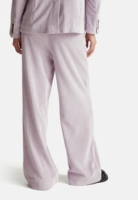 Jasnofioletowe szerokie spodnie z korduroyu z pionowymi prążkami, o luźnym kroju i gładkiej fakturze. Widać czarne obuwie.