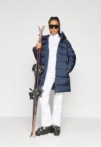 Cappotto imbottito blu navy con cappuccio, maglione bianco con zip, pantaloni da sci bianchi, scarponi da sci neri e un paio di sci. Gli occhiali da sole completano l'outfit per gli sport invernali.