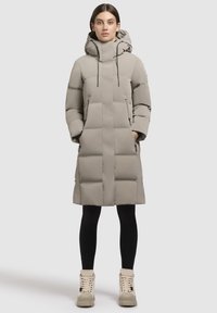 Bēšs pūkains mētelis ar kapuci, kam ir izšūts dizains, divas priekšējās kabatas un melni auklas. Uzvilkts virs melnām zeķubiksēm un masīva dizaina bēšām zābakiem.