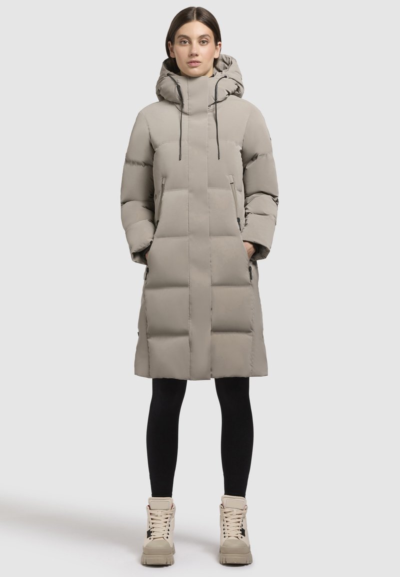 Bēšs pūkains mētelis ar kapuci, kam ir izšūts dizains, divas priekšējās kabatas un melni auklas. Uzvilkts virs melnām zeķubiksēm un masīva dizaina bēšām zābakiem.