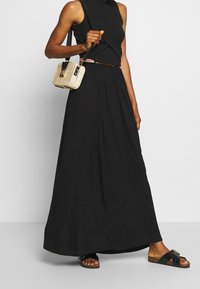 Top negro sin mangas con detalle retorcido, falda maxi negra fluida, sandalias negras y un pequeño bolso cruzado de tejido con detalles en oro.