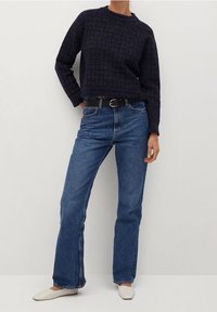 Pull texturé bleu marine à manches longues, associé à un jean bleu coupe droite et une ceinture noire. Des chaussures blanches à bout pointu complètent le look.