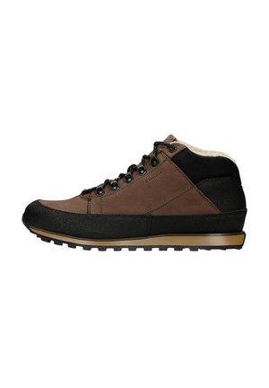 Personne portant un pantalon noir et des chaussures de randonnée marron et noires à semelles texturées, posant le pied sur une surface blanche, vue de côté et de dos.