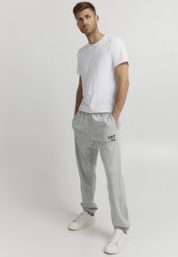 Hvit t-skjorte med kort erme, parret med lysegrå joggebukser med svart trykk av "EST. '99". Modellen har på seg hvite joggesko.