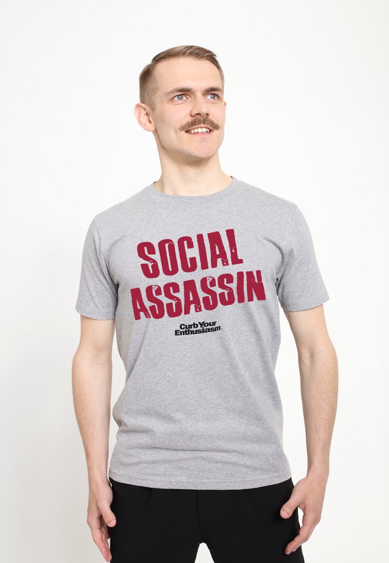 Camiseta de algodón gris con mangas cortas que presenta un texto rojo en negrita "SOCIAL ASSASSIN" y un texto negro más pequeño "Curb Your Enthusiasm". Diseño sencillo.