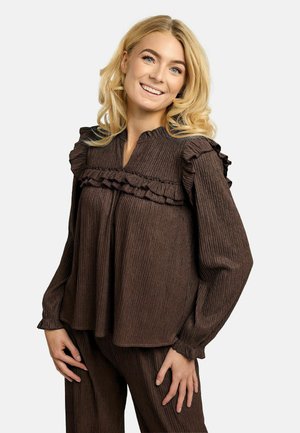 Naeratav blondide juustega naine, kes kannab pruuni tekstuuriga pika varrukaga pluusi rüüsidega ja sobivaid pükse, seisab ühevärvilise tausta ees.