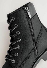 Schwarze Leder-Ankle-Boots mit runden Schnürsenkeln, Metallösen, seitlichem Reißverschluss und gepolstertem Schaft vor einem weißen Hintergrund.