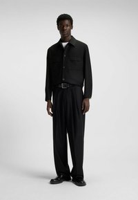 Zwarte button-up jas met twee borstzakken, gecombineerd met losse zwarte broeken en zwarte schoenen. Soepele textuur en modern ontwerp.