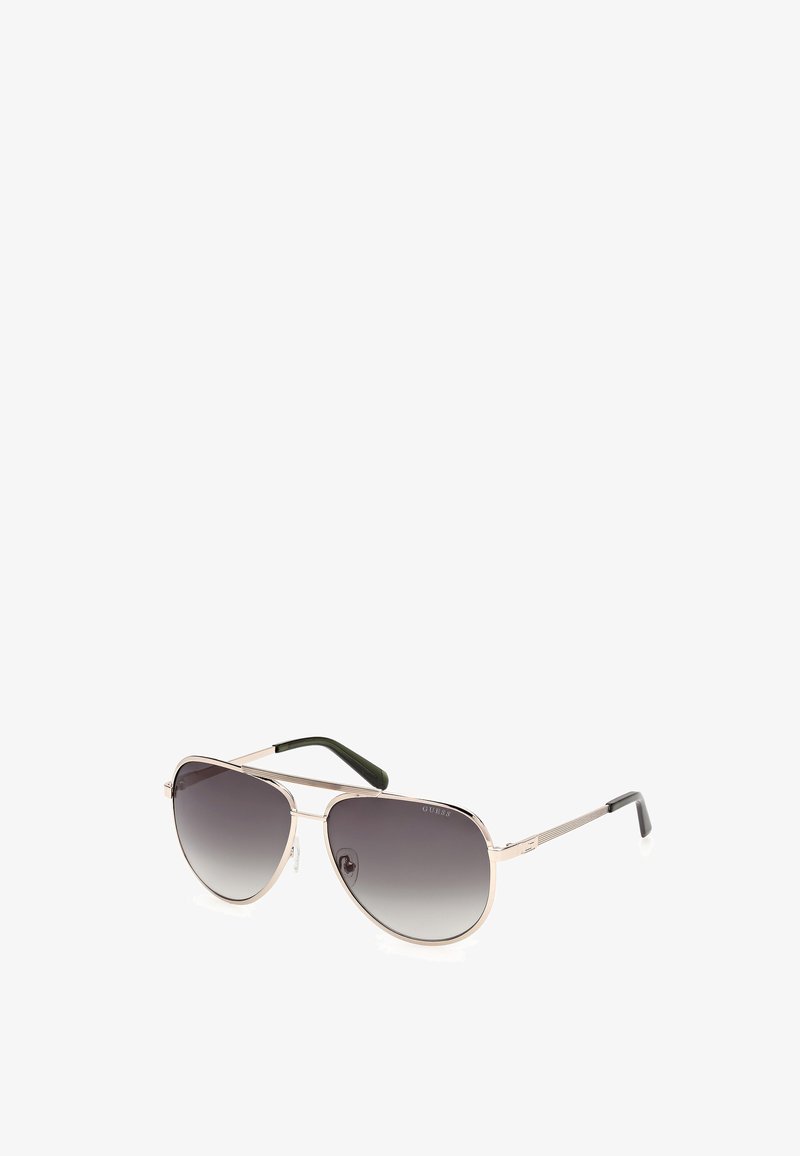 Óculos de sol estilo aviador com lentes cinza em degradê, armação de metal em tom dourado e detalhes em borracha verde nas hastes. Hastes finas com detalhe texturizado.