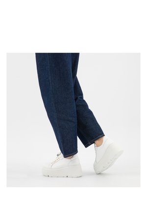 Persona che indossa jeans blu scuro e sneakers bianche con piattaforma, con un piede leggermente sollevato su sfondo bianco uniforme.