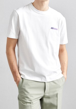 Mann trägt ein schlichtes weißes kurzärmliges T-Shirt mit kleinem lila Logo auf der linken Brust, kombiniert mit hellbeigen Hosen.