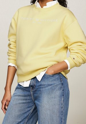 Osoba nosząca bladożółtą bluzę Tommy Hilfiger na białej koszuli i niebieskich dżinsach, z ręką w kieszeni, logo marki widoczne na piersi i rękawie.