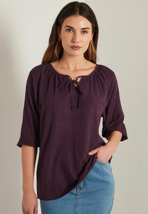 Blusa viola, ampia e morbida, con dettaglio a fiocco sul collo. Il design presenta maniche a tre quarti e un tessuto morbido e textured.