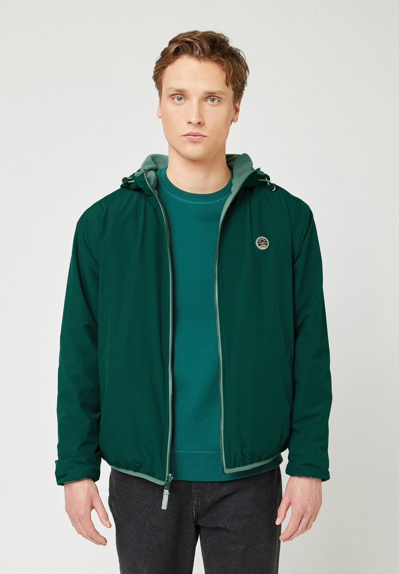 Giacca verde con zip e cappuccio, con polsini elastici, indossata sopra una maglietta verde abbinata. Presenta un logo rotondo sul lato sinistro del petto.