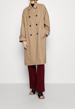 Personne portant un long manteau beige croisé, un pantalon bordeaux et des chaussures à talons ouvertes noires, debout contre un mur blanc.