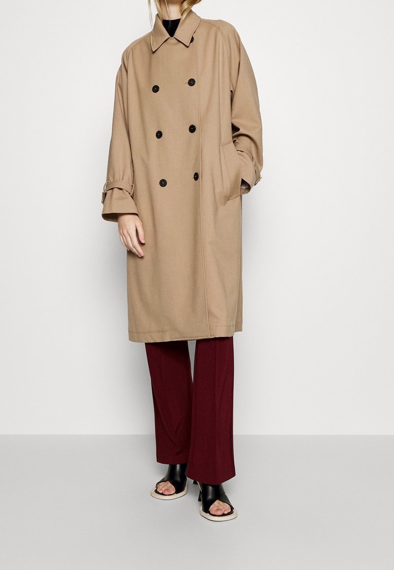 Personne portant un long manteau beige croisé, un pantalon bordeaux et des chaussures à talons ouvertes noires, debout contre un mur blanc.