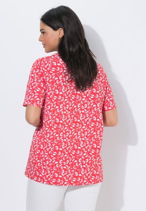 Donna con capelli scuri che indossa una maglietta rossa a maniche corte con motivo floreale bianco, vista da dietro, abbinata a pantaloni bianchi.