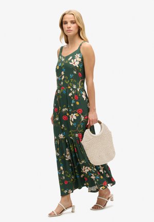 Maxi sukienka w ciemnej zieleni, z czerwonymi i niebieskimi kwiatami, dopasowana góra, rozszerzany dół, w połączeniu z tkanym beżowym torbą i białymi sandałami.