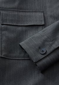 Grijs gestreept stof met een borstzak en knoopdetail op de manchetten. Het materiaal heeft een subtiele textuur en een getailleerd ontwerp.