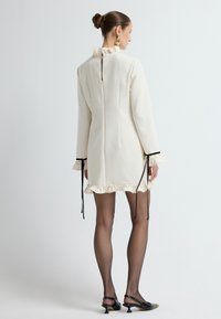 Abito color crema con accenti a volant sulle maniche e sull'orlo, accompagnato da lacci neri, con chiusura a zip sul retro.