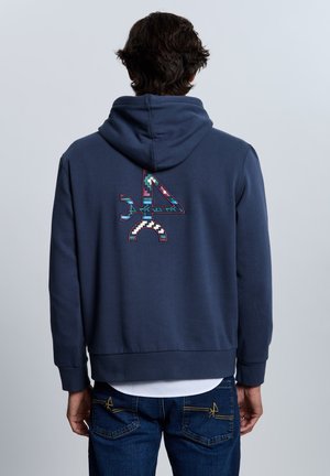 Hombre vestido con sudadera azul marino con logo geométrico 4 colorido en la espalda, combinado con vaqueros azules, de pie frente a un fondo gris claro.