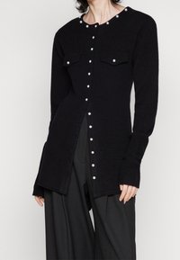 Mujer vistiendo un cárdigan negro de manga larga con botones de perla y dos bolsillos en el pecho, combinado con pantalones anchos negros.