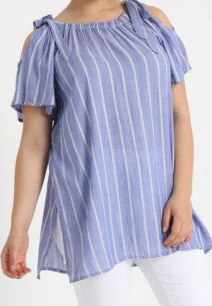 Kobieta ubrana w luźną bluzkę z odkrytymi ramionami w pionowe paski w jasnoniebieskim i białym kolorze z wiązanymi ramiączkami oraz białe spodnie.