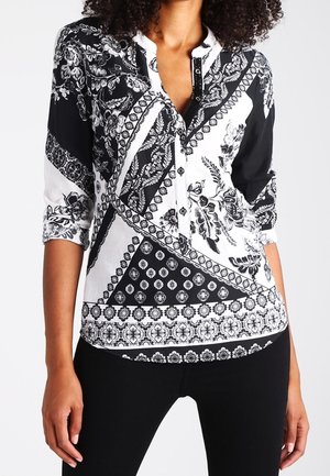 Mujer con blusa de estampado en blanco y negro con diseños florales y geométricos y pantalones negros, brazos relajados a los lados.