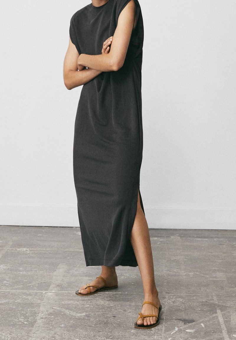 Höyhenlakan, tummanharmaa maksi mekko, jossa on lyhyet hihat ja sivuhalkiot. Yhdistetty ruskeisiin litteisiin sandaaleihin, harmaasta teksturoidusta lattia-taustasta.