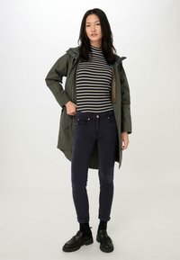 Olijfgroene waterdichte jas met capuchon over een zwart-wit gestreepte coltrui, gecombineerd met donkerblauwe skinny jeans en zwarte ballerina's.
