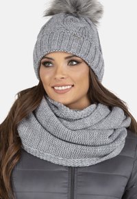 Strikket grå beanie med en pom-pom og tørklæde; begge har et struktureret, chunky mønster, parret med en sort, polstret jakke.