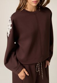 Bruine sweatshirt met ronde hals en lange mouwen, voorzien van witte bloemenborduurwerk op één schouder en een trekkoord in de taille.