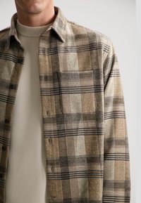 Cercel de cămașă cu carouri maro, cu dungi de culoare albastră închis și negru, având un buzunar, închidere cu nasturi și un material moale, texturat. Purtați deasupra unui pulover crem.
