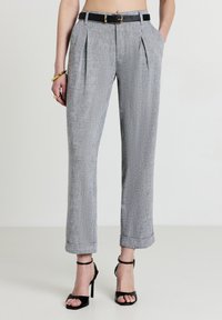 Donna che indossa pantaloni cropped grigi plissettati con una cintura nera, gioielli dorati e sandali neri con tacco alto, in piedi su un pavimento bianco.