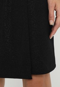 Zwarte jurk met een gestructureerde afwerking en subtiele glitter. Het heeft een rechte snit met een kleine zijsplit voor beweging.