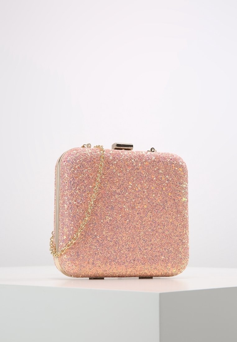 Petit sac à main carré rose pailleté avec une bandoulière en chaîne dorée, placé sur un socle blanc sur fond blanc uni.