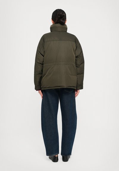 Person with dark hair in a bun wearing a dark green puffer jacket, wide-leg dark blue jeans, and black shoes, standing facing away.