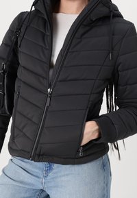 Czarna, pikowana kurtka z zamkiem z przodu, przypinanym kapturem i ściągaczami; ma ukośny wzór pikowania i boczne kieszenie.