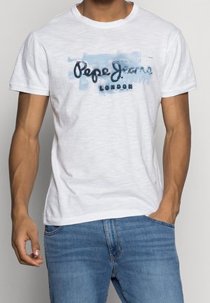 Man in een wit Pepe Jeans London t-shirt en blauwe jeans, met één hand in zijn zak en de andere ontspannen naast zich.