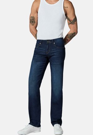 Mann trägt dunkelblaue Jeans mit geradem Bein, weißes Tanktop, weiße Sneakers, mit tätowierten Armen sichtbar vor einfachem Hintergrund.