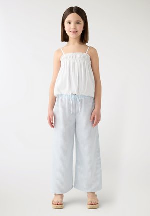 Dziewczyna z prostymi brązowymi włosami ubrana w biały top bez rękawów, jasnoniebieskie szerokie spodnie w paski i beżowe sandały na platformie, stoi na białym tle.